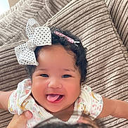 Magdalene is registered to the contest to win money with this photo: baby, child, smiling, tongue_out, bow, hair_accessory, curly_hair, infant, cute, happy, person, portrait, indoor, couch, clothing, hand, playful, face, skin, young