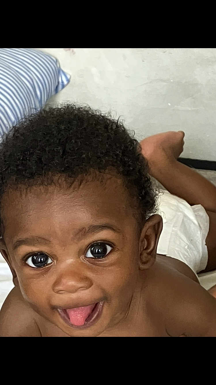 Kyzier Williams joined the competition — help win amazing prizes! baby, child, curly_hair, tongue_out, smiling, face, eyes, skin, bed, pillow, diaper, happy, cute, infant, lying_down, closeup, person, indoors, playful, young_child