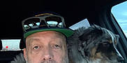 Bru is registered to the contest to win money with this photo: man, dog, car, cap, sunglasses, shoulder, seatbelt, tattoo, beard, white_tank_top, pet, animal, window, sky, interior, portrait, casual, adult, companion, travel