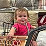 baby, child, shopping_cart, pink_clothing, tongue_out, pillow, blanket, store, retail, shelf, home_decor, foot, hand, smile, cute, infant, seat, pattern, fabric, aisle