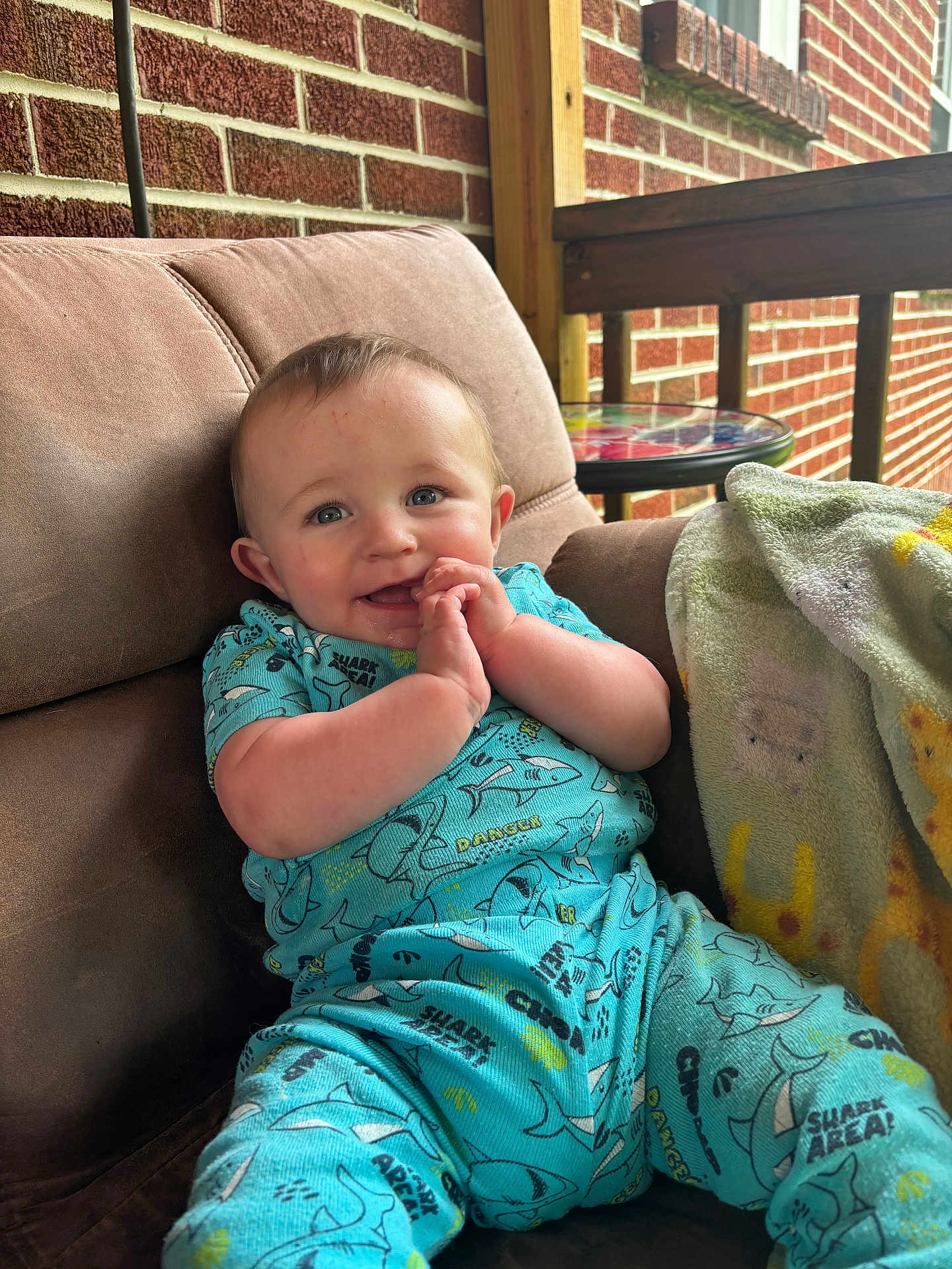 Mason is registered to the contest to win money with this photo: baby, child, smiling, blue_outfit, shark_pattern, cushioned_chair, blanket, brick_wall, porch, wooden_railing, happy, face, hands, sitting, indoor, cute, infant, clothing, comfort, portrait