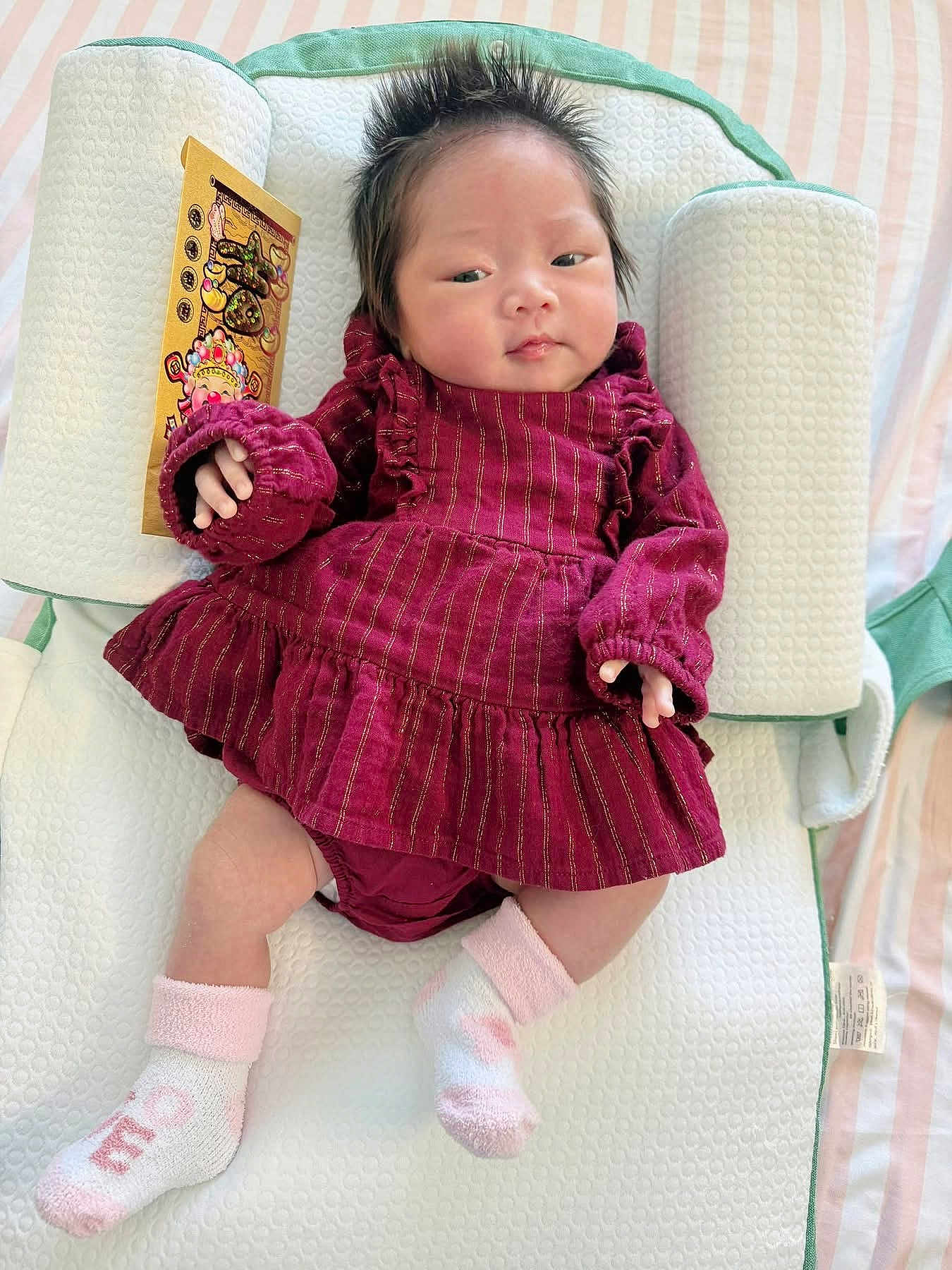 Giana is registered to the contest to win money with this photo: baby, infant, newborn, child, maroon_dress, socks, baby_sock, pillow, baby_lounger, hair, tufted_hair, red_envelope, hand, face, eyes, blanket, striped_sheet, portrait, laying_down, cute