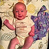 baby, infant, smile, onesie, quilt, seashell_pattern, clothing, purple_garment, happy, lying_down, cute, newborn, celebration, indoor, child, fabric, pattern, toddler, person, portrait