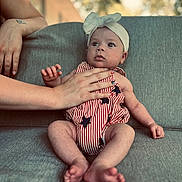 Esmae joined the competition — help win amazing prizes! baby, infant, headband, bow, striped_clothing, stars, hand, couch, gray_couch, sitting, person, adult_hand, outdoor_background, blurred_background, natural_light, portrait, cute, young_child, skin, expression