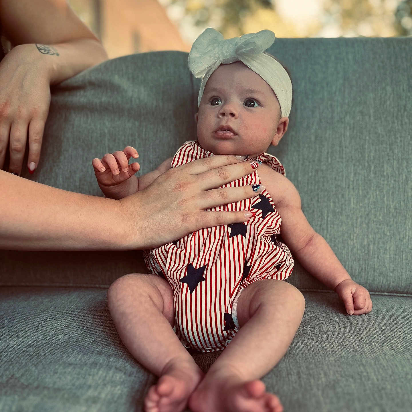 Esmae joined the competition — help win amazing prizes! adult_hand, baby, blurred_background, bow, couch, cute, expression, gray_couch, hand, headband, infant, natural_light, outdoor_background, person, portrait, sitting, skin, stars, striped_clothing, young_child