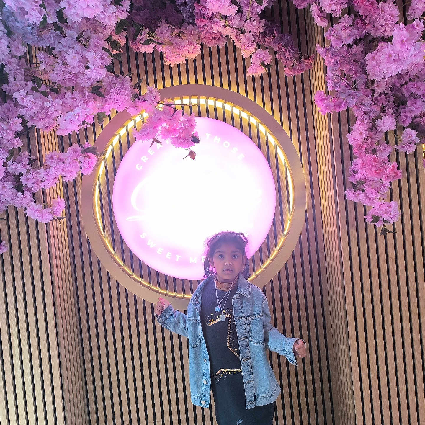 Muqadas joined the competition — help win amazing prizes! blossoms, boots, casual_clothing, child, curly_hair, decor, denim_jacket, fashion, floor, girl, glowing_sign, indoor, lighting, necklaces, pink_flowers, portrait, standing, vertical_slats, wooden_wall, young_person