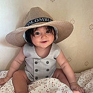 Naim is registered to the contest to win money with this photo: baby, child, hat, smile, happy, indoors, blanket, floral_pattern, sitting, cute, portrait, clothing, infant, face, skin, person, young, headwear, buttoned_outfit, room