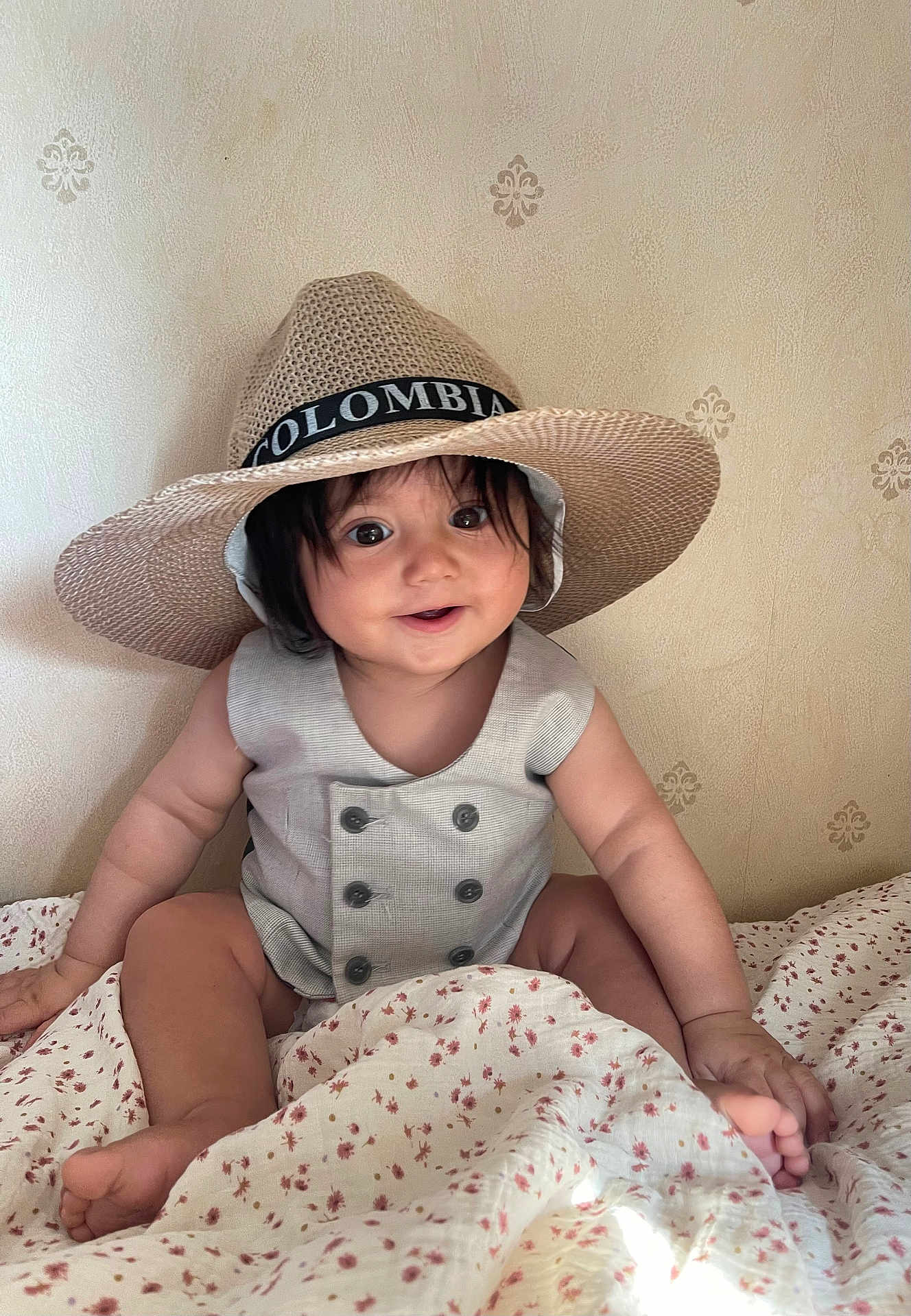 Naim is registered to the contest to win money with this photo: baby, child, hat, smile, happy, indoors, blanket, floral_pattern, sitting, cute, portrait, clothing, infant, face, skin, person, young, headwear, buttoned_outfit, room