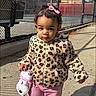 child, toddler, girl, outdoor, sidewalk, leopard_print_coat, pink_pants, pink_shoes, hair_bow, curly_hair, stuffed_animal, unicorn_toy, walking, smiling, portrait, sports_court, chain_link_fence, sunlight, shadows, street_style