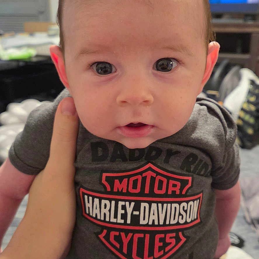 Gunner is registered to the contest to win money with this photo: baby, infant, child, mohawk, harley_davidson, onesie, clothing, hand, indoor, furniture, television, person, cute, portrait, looking_at_camera, skin, face, eyes, sitting, babywear