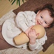 Leya a rejoint le concours — aidez-le/la à gagner de superbes lots ! toddler, child, smiling, blue_eyes, pigtails, white_sweater, brown_leggings, wooden_doll, blanket, pillow, flower, indoor, cozy, cute, happy, portrait, soft_texture, laying_down, toy, decor