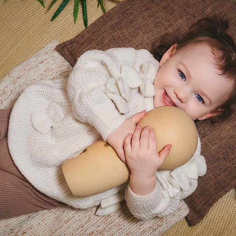 Leya a rejoint le concours — aidez-le/la à gagner de superbes lots ! toddler, child, smiling, blue_eyes, pigtails, white_sweater, brown_leggings, wooden_doll, blanket, pillow, flower, indoor, cozy, cute, happy, portrait, soft_texture, laying_down, toy, decor