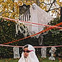 child, ghost_costume, sunglasses, halloween, autumn, tombstones, decorations, ghost, caution_tape, grass, tree, leaves, spooky, outdoor, holiday, costume, fun, playful, fall, yard