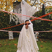Austin is registered to the contest to win money with this photo: child, costume, ghost, sunglasses, halloween, yard, grass, fall_leaves, decoration, tombstone, caution_tape, spooky, outdoor, tree, shrubbery, holiday, fun, playful, white_sheet, face
