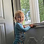 toddler, child, pajamas, door, window, screen, indoors, natural_light, curly_hair, blue_clothing, standing, face, expression, hand, home, background, greenery, daylight, portrait, person
