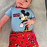 Julius is registered to the contest to win money with this photo: baby, child, mickey_mouse, clothing, red_pants, blue_shirt, lying_down, cute, smiling, infant, patterned_pants, cartoon_character, soft_surface, portrait, casual, young_child, hand_gesture, bright_colors, comfortable, indoors