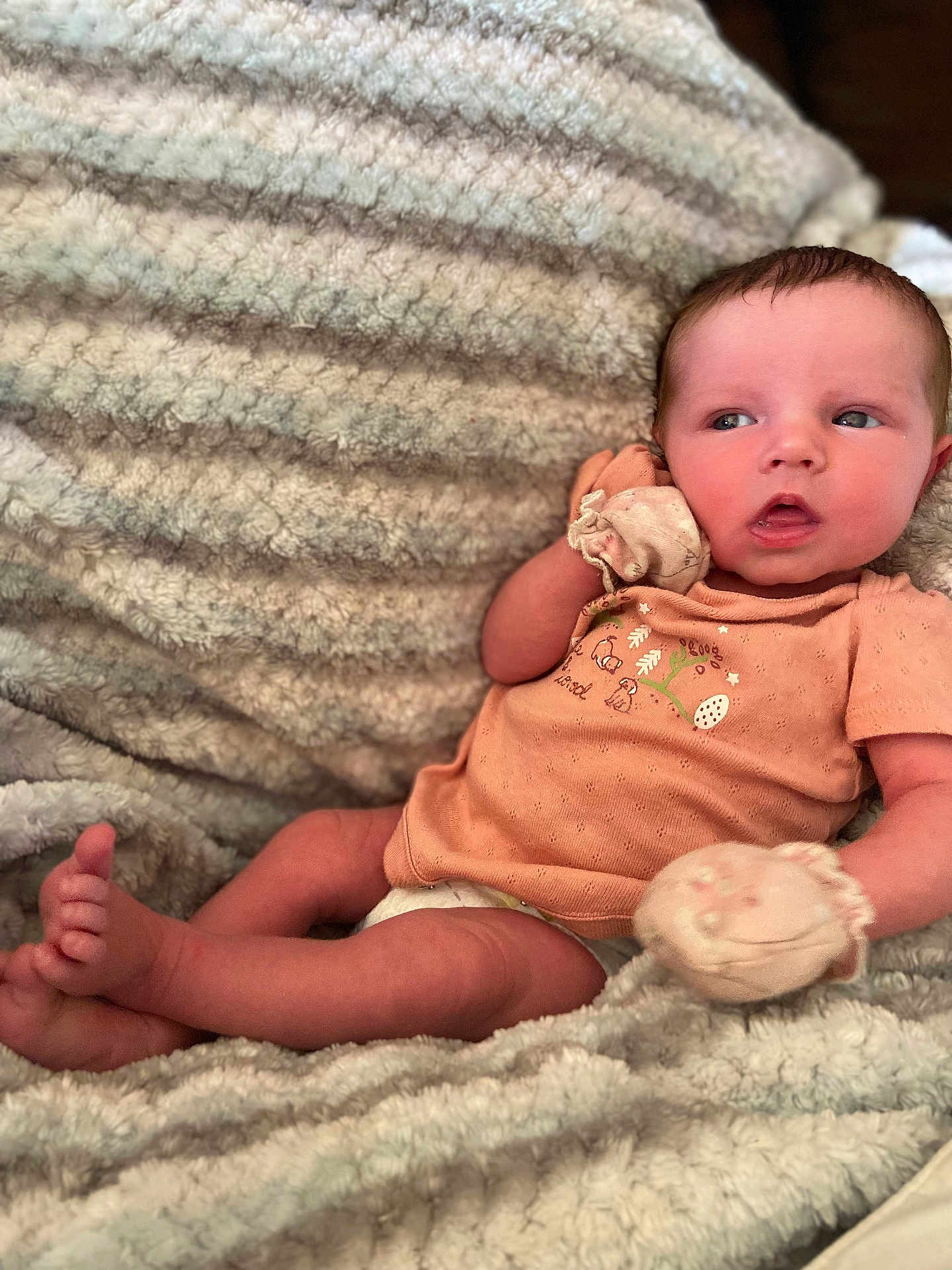 April is registered to the contest to win money with this photo: baby, infant, blanket, mittens, clothing, person, face, legs, feet, lying_down, soft_texture, striped_blanket, peach_shirt, cute, newborn, resting, indoors, skin, expression, portrait