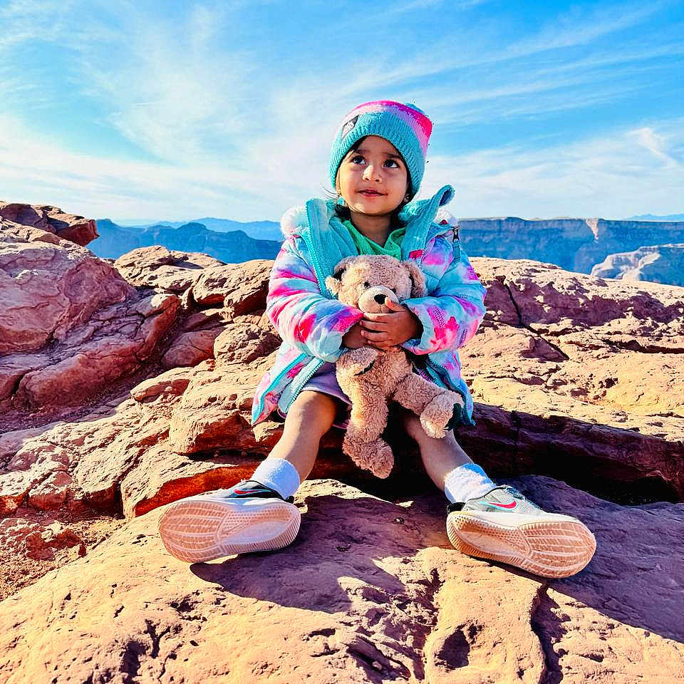 Aarya is registered to the contest to win money with this photo: child, clothing, coat, face, female, footwear, girl, head, nature, outdoors, person, photography, portrait, rock, shoe, sitting, sky, teddybear, toy, wilderness
