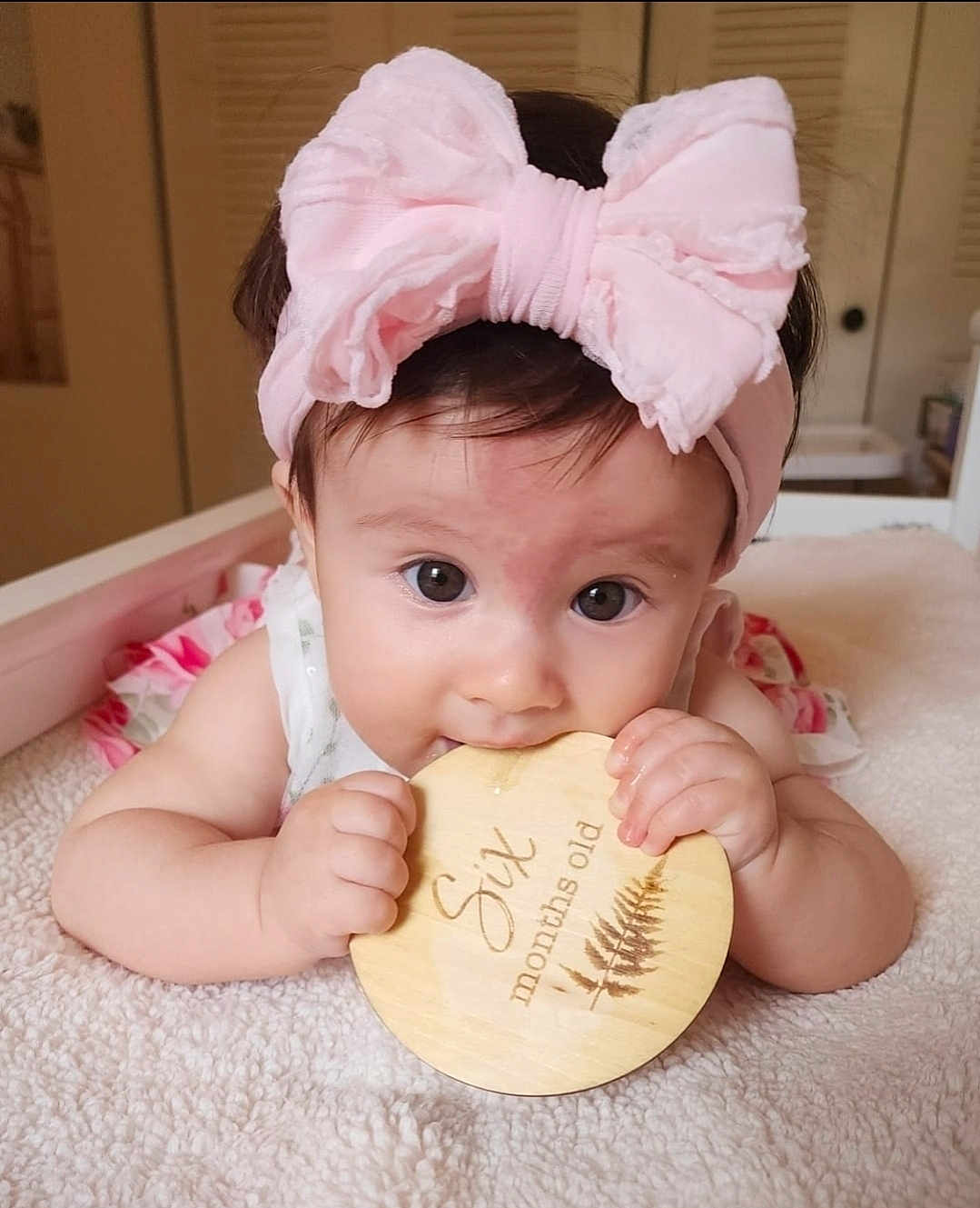 Camila is registered to the contest to win money with this photo: baby, infant, headband, pink_bow, wooden_plaque, chewing, milestone, six_months_old, blanket, cute, child, portrait, indoors, soft_texture, hands, face, big_eyes, lying_down, baby_clothes, home