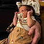 baby, newborn, infant, pacifier, pacifier_clip, onesie, patterned_pants, adult_hand, blanket, couch, close_up, portrait, indoor, soft_texture, warm_lighting, looking_up, cute, family_moment, tiny_hands, brown_clothing
