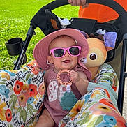 Hadley joined the competition — help win amazing prizes! baby, child, sunglasses, hat, stroller, toy, hand, person, outdoor, greenery, grass, sunlight, floral_pattern, colorful, happy, smiling, summer, casual_clothing, infant, playful