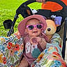 baby, child, sunglasses, hat, stroller, toy, hand, person, outdoor, greenery, grass, sunlight, floral_pattern, colorful, happy, smiling, summer, casual_clothing, infant, playful