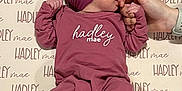 Hadley is registered to the contest to win money with this photo: baby, infant, child, hands, clothing, headband, foot, background, pattern, name, sucking, cute, pink, mauve, person, newborn, fabric, laying, holding, portrait
