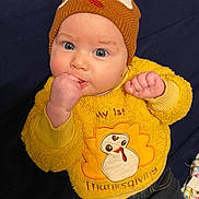 Journey is registered to the contest to win money with this photo: baby, child, blue_eyes, knit_hat, yellow_sweater, thanksgiving, turkey, cute, infant, fist_in_mouth, clothing, dark_background, portrait, indoors, cozy, cute_hat, happy, person, toddler, sitting