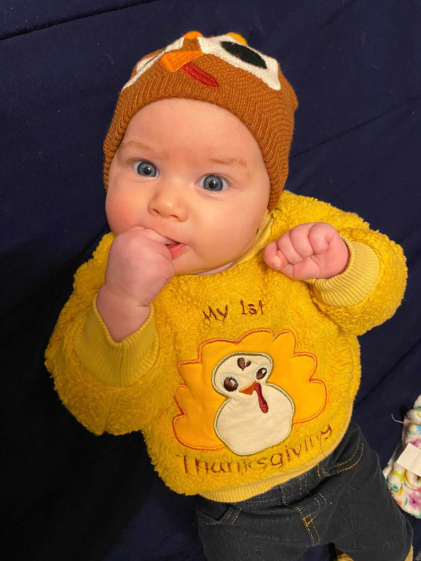 Journey is registered to the contest to win money with this photo: baby, child, blue_eyes, knit_hat, yellow_sweater, thanksgiving, turkey, cute, infant, fist_in_mouth, clothing, dark_background, portrait, indoors, cozy, cute_hat, happy, person, toddler, sitting