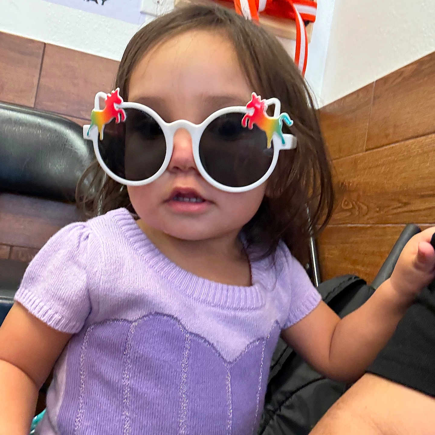 Amelia is registered to the contest to win money with this photo: accessory, child, colorful, curious, cute, fashion, fun, indoor, person, playful, portrait, purple_top, seated, short_hair, sunglasses, tattoo, toddler, unicorn, wood_paneling, young
