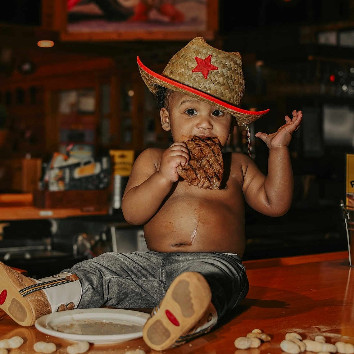 Jiayze is registered to the contest to win money with this photo: baby, bare_chest, boots, casual, child, cowboy_hat, cute, eating, food, indoor, mischievous, pants, peanuts, person, portrait, restaurant, snack, steak, toddler, wooden_counter