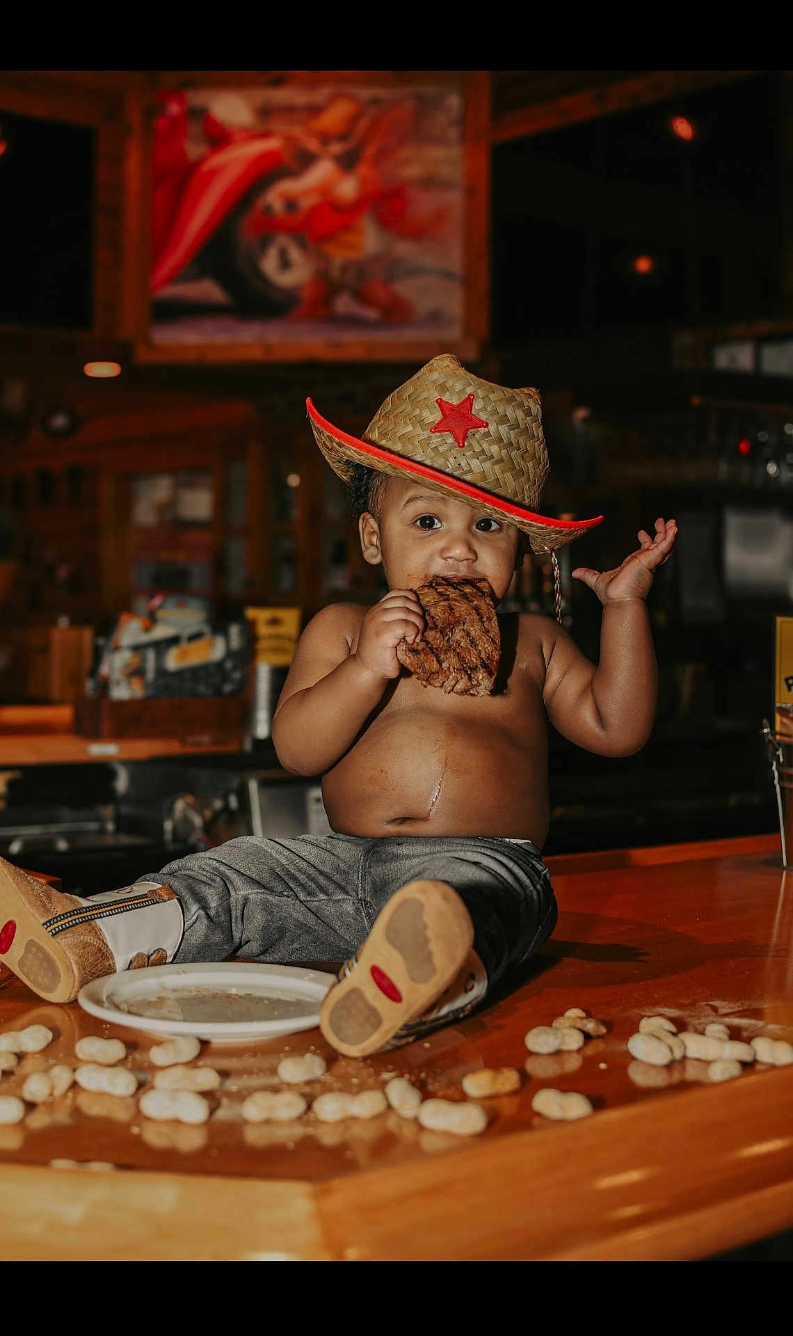 Jiayze is registered to the contest to win money with this photo: toddler, child, cowboy_hat, steak, eating, bare_chest, pants, boots, peanuts, wooden_counter, indoor, restaurant, food, snack, cute, mischievous, portrait, baby, person, casual