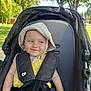 baby, child, stroller, hat, yellow_outfit, smile, outdoor, park, sunlight, greenery, trees, safety_harness, cute, infant, seat, summer, expression, person, daylight, casual