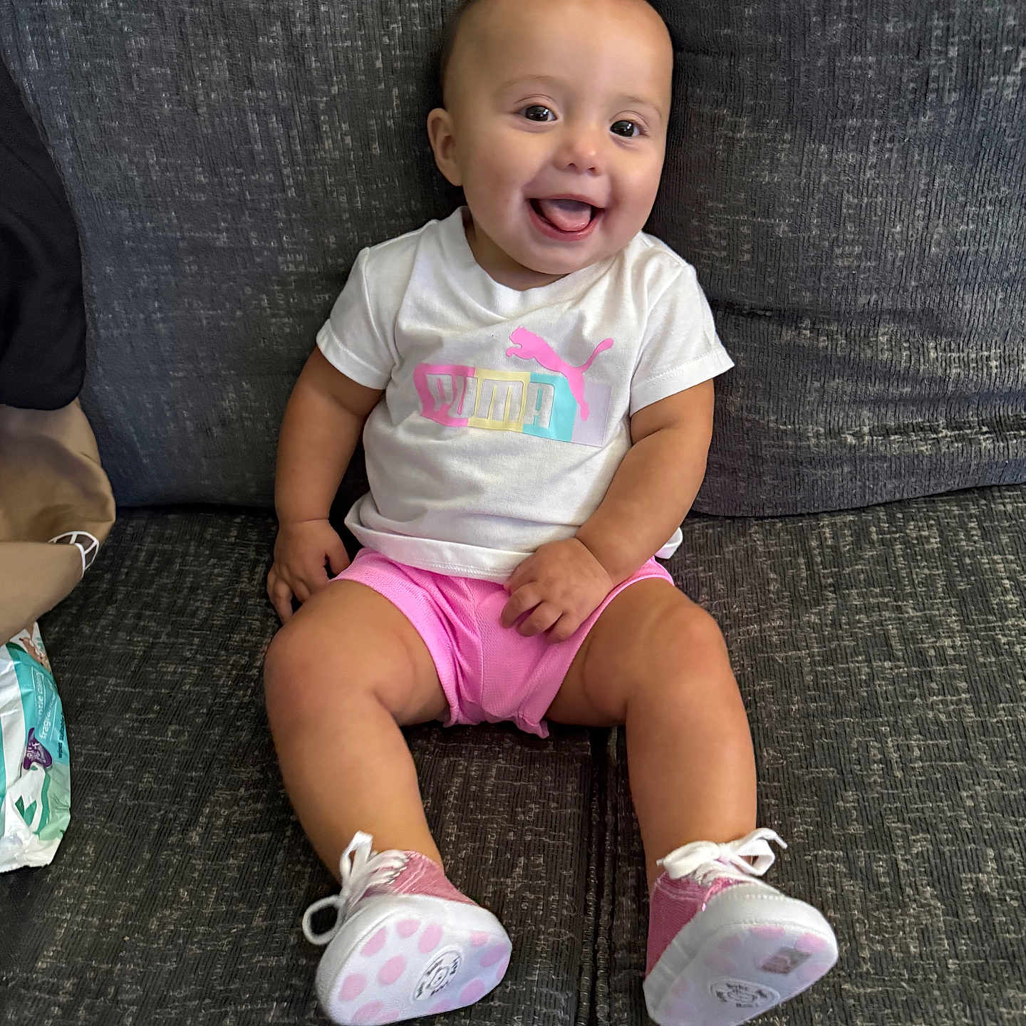Eliana is registered to the contest to win money with this photo: apparel, baby, child, clothing, couch, cute, face, footwear, happy, indoor, infant, person, pink, seated, shoes, shorts, smiling, tongue, tshirt, white