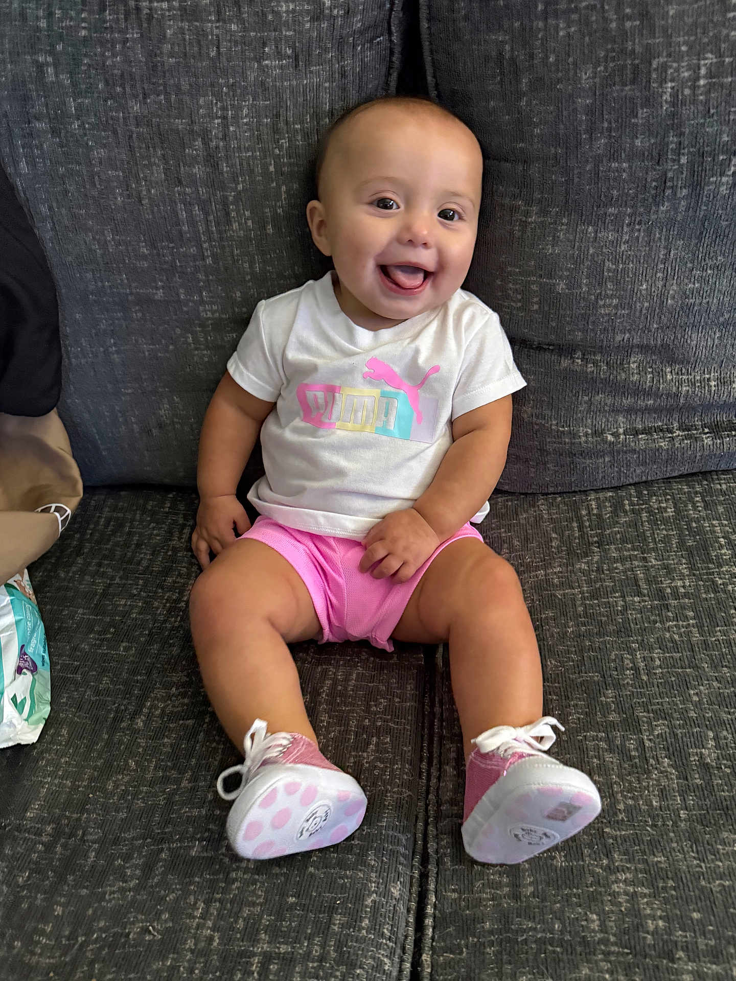Eliana is registered to the contest to win money with this photo: baby, child, smiling, happy, couch, clothing, tshirt, shorts, shoes, footwear, person, indoor, seated, pink, white, face, tongue, cute, infant, apparel