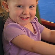 Melody is registered to the contest to win money with this photo: child, toddler, smiling, purple_shirt, blonde_hair, blue_eyes, seat, black_shorts, happy, portrait, indoor, person, cute, young_child, casual_clothing, fun, joy, arm, hair, face