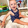 toddler, child, smiling, blue_eyes, romper, toy_boat, white_flower, indoor, wooden_floor, baby_walker, face, happy, sitting, decor, vase, red_bracelet, furniture, person, playful, expression