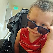 Jakob is registered to the contest to win money with this photo: toddler, child, sunglasses, stroller, indoor, red_shirt, seatbelt, britax, light, exit_sign, hallway, curious, person, young_child, casual, short_hair, baby, portrait, baby_carrier, expression