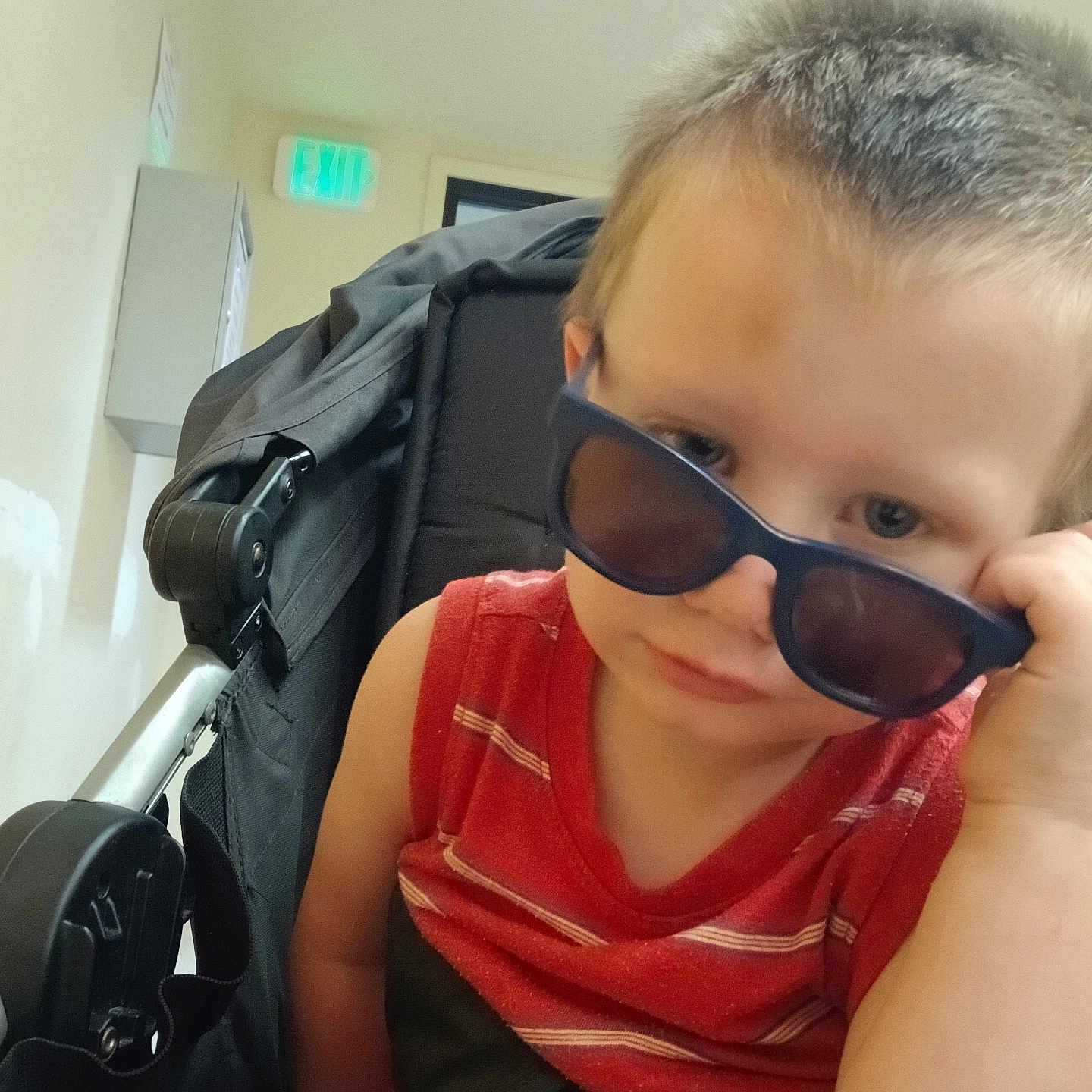 Jakob is registered to the contest to win money with this photo: baby, baby_carrier, britax, casual, child, curious, exit_sign, expression, hallway, indoor, light, person, portrait, red_shirt, seatbelt, short_hair, stroller, sunglasses, toddler, young_child