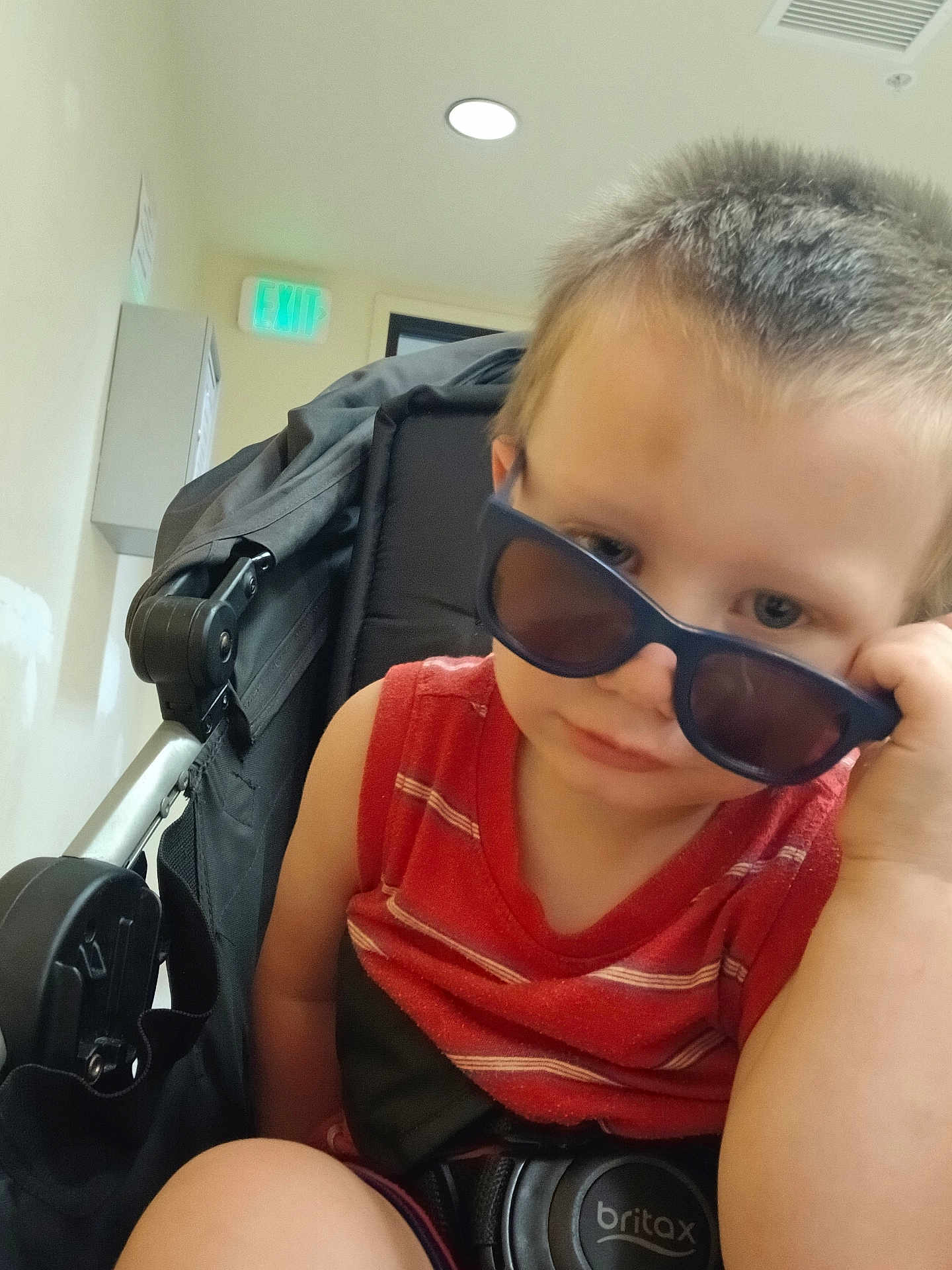 Jakob is registered to the contest to win money with this photo: toddler, child, sunglasses, stroller, indoor, red_shirt, seatbelt, britax, light, exit_sign, hallway, curious, person, young_child, casual, short_hair, baby, portrait, baby_carrier, expression