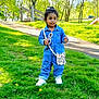 child, toddler, grass, park, trees, sunlight, denim_jumpsuit, white_sneakers, sunglasses, purse, outdoor, walking_path, fence, greenery, daylight, person, nature, casual_clothing, cute, playful