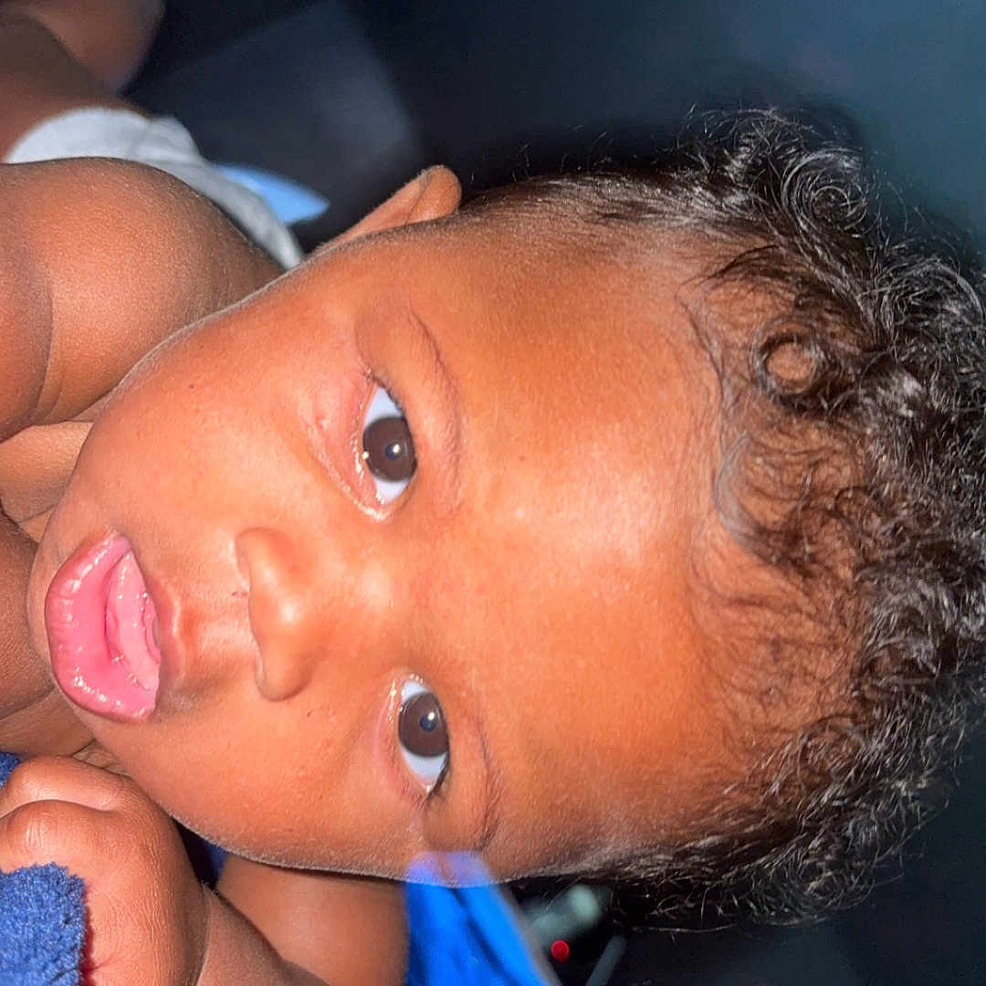 Kamora is registered to the contest to win money with this photo: baby, blanket, blue_blanket, brown_eyes, child, close_up, curly_hair, cute, expression, face, hand, indoor, infant, laying_down, looking, person, portrait, skin, skin_tone, young_child