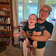 Hannah is registered to the contest to win money with this photo: baby, infant, adult, woman, smiling, happy, portrait, indoors, home, family, holding, arms, bookshelf, books, toy, window, kitchen, clothing, glasses, cap