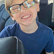 Aiden is registered to the contest to win money with this photo: child, boy, glasses, smile, car_interior, seat, shorts, blue_shirt, reflection, sunlight, window, vehicle, casual_clothing, happy, young, portrait, sitting, daylight, transport, backseat