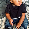 toddler, child, afro_hair, black_shirt, ripped_jeans, sneakers, bedspread, striped_bedspread, blue_ball, indoor, curious_expression, seated, hands, young_child, casual_clothing, footwear, portrait, home, person, looking_away
