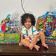 Manny joined the competition — help win amazing prizes! child, curly_hair, smiling, striped_shirt, toys, toy_cars, packaging, wooden_surface, indoor, colorful, happy, sitting, playful, wrapped_toys, plastic_wrap, floor, young_child, person, casual_clothing, portrait