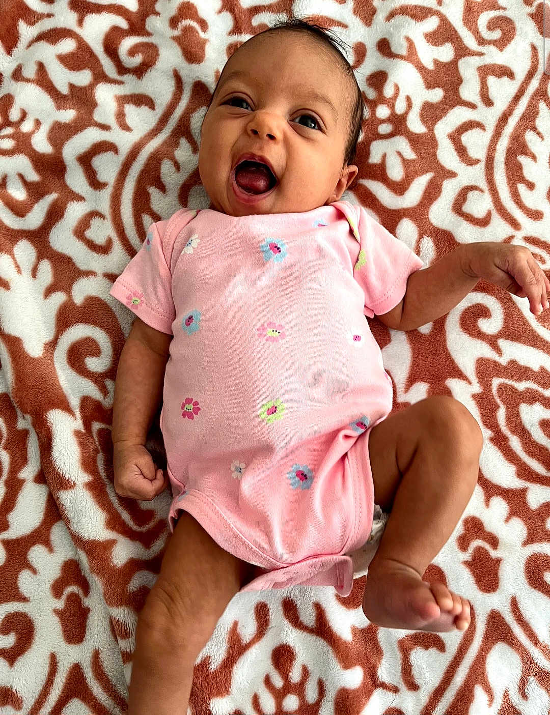Luna is registered to the contest to win money with this photo: baby, infant, child, smiling, onesie, pink_clothing, blanket, patterned_blanket, happy, lying_down, cute, human, skin, face, expression, small_hand, small_foot, indoors, soft_texture, portrait