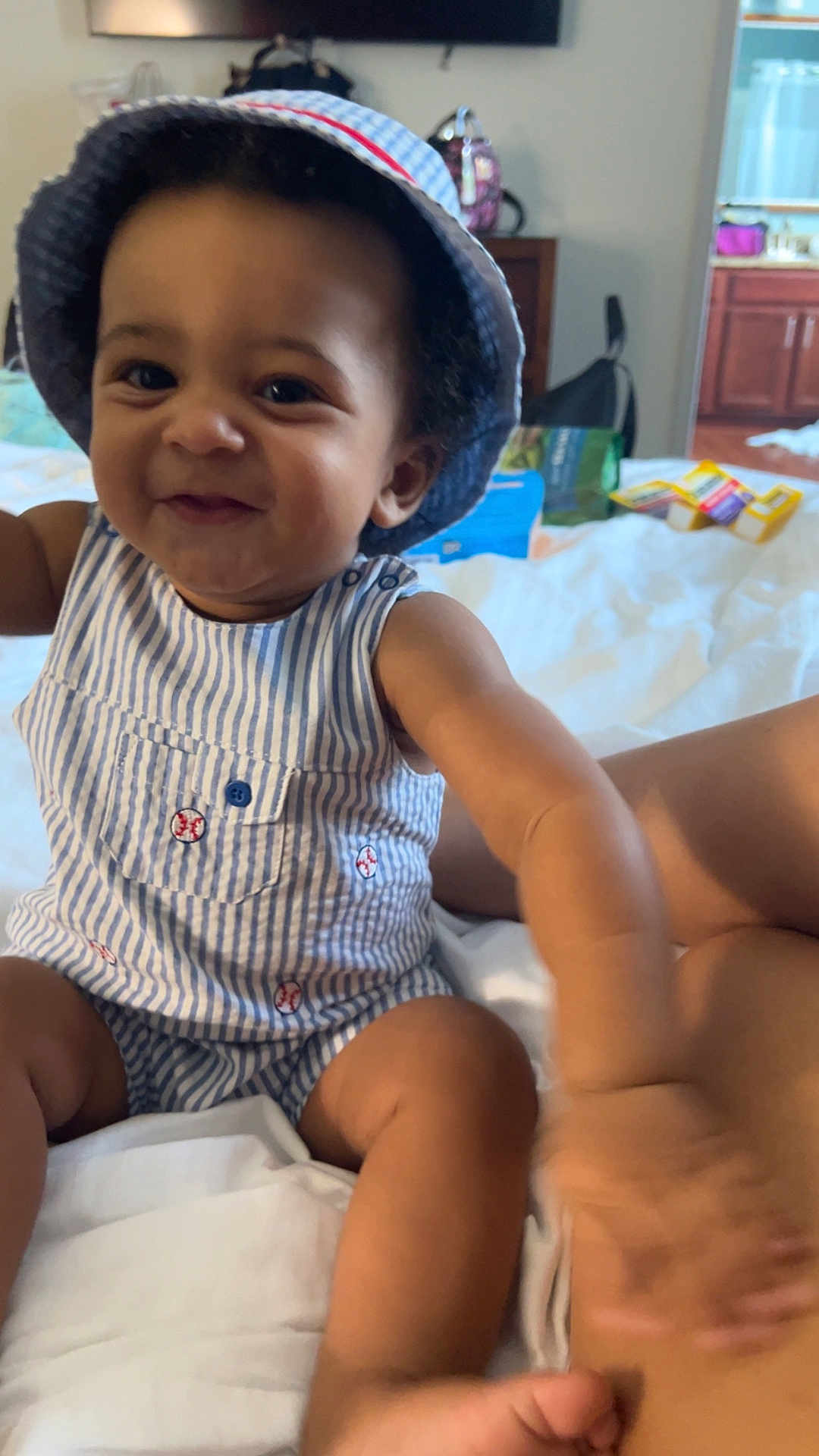 Leo is registered to the contest to win money with this photo: baby, child, smiling, hat, striped_clothing, romper, bed, indoor, person, cute, happy, skin, face, arm, hand, foot, room, furniture, blurred_background, playful