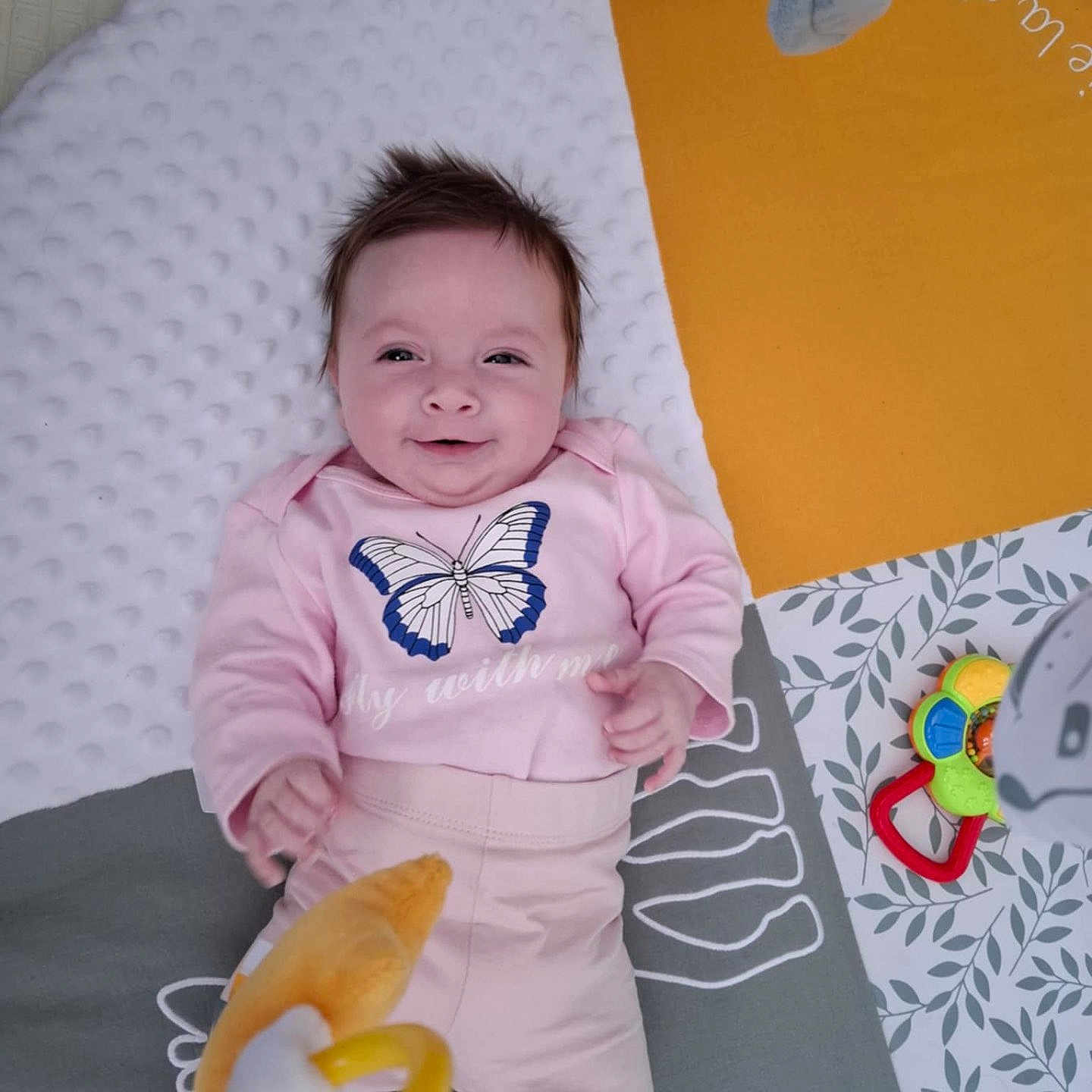 Luna a rejoint le concours — aidez-le/la à gagner de superbes lots ! baby, butterfly_print, child, cute, face, footwear, happy, indoor, infant, lying_down, patterned_mat, person, pink_clothing, play_mat, playtime, smiling, sock, soft_texture, toy, yellow_ring