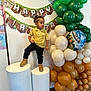 toddler, child, birthday, party, balloons, minecraft, yellow_shirt, black_pants, boots, banner, indoor, celebration, decorations, standing, pedestal, smiling, festive, face, person, event